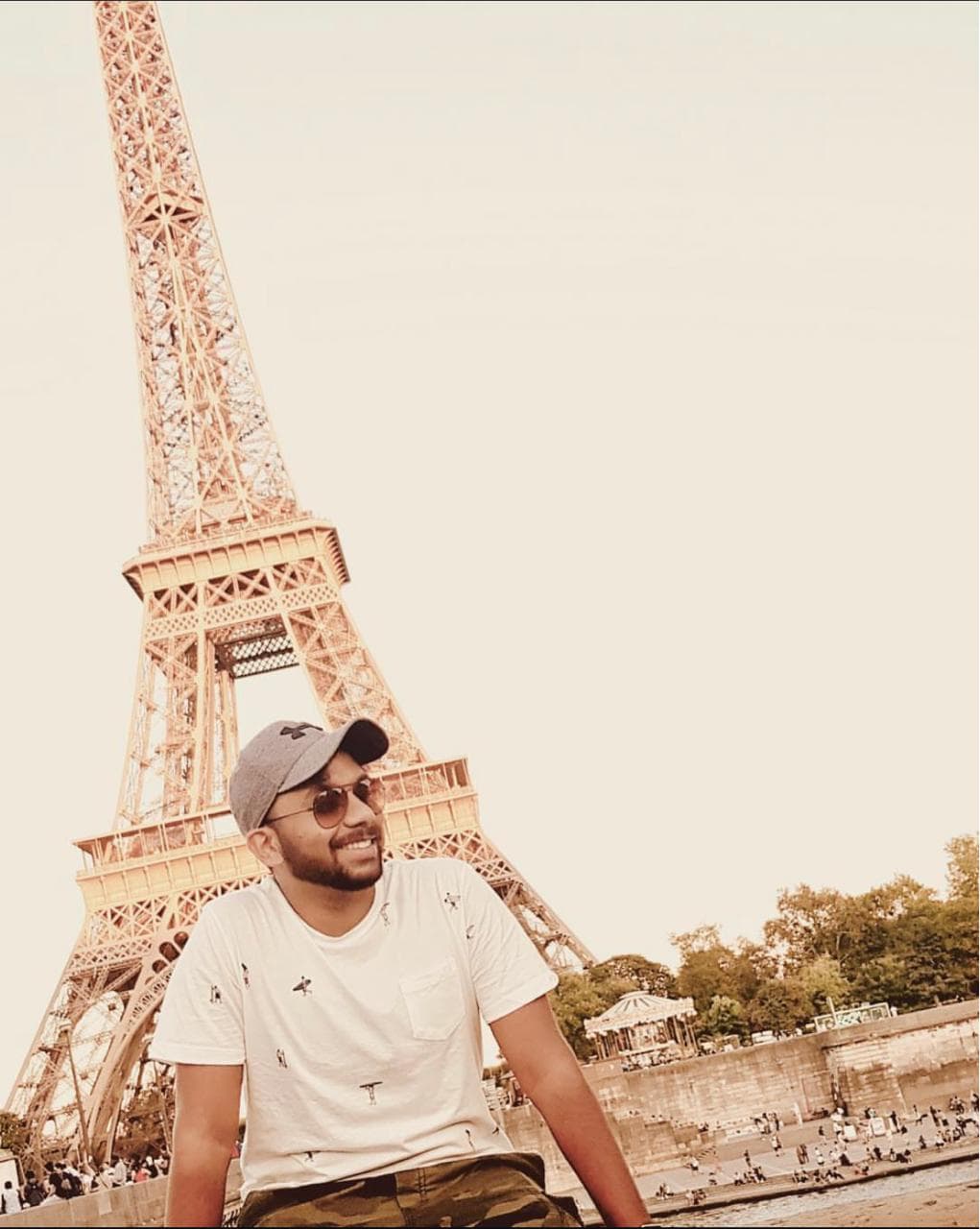 Smiling man in a cap and sunglasses posing in front of the Eiffel Tower.